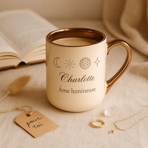 Può includere: Tazza in ceramica color crema con finiture dorate, piena di una bevanda. La tazza presenta simboli celesti e il nome "Charlotte" con "Âme lumineuse" sotto. Un'etichetta con "pour toi" è allegata.
