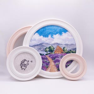 May include: A collection of framed cross-stitch art. The largest frame features a landscape with a house, lavender field, and blue sky. Other frames include a hedgehog and a pink frame. The frames are white and pink.
