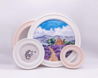 May include: A collection of framed cross-stitch art. The largest frame features a landscape with a house, lavender field, and blue sky. Other frames include a hedgehog and a pink frame. The frames are white and pink.