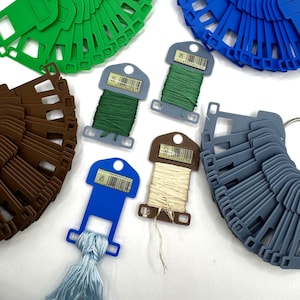 May include: A collection of plastic embroidery floss bobbins in green, blue, brown, and gray, arranged in circular fan-like clusters. Several individual bobbins are wrapped with thread in shades of green, light blue, and cream, each with a barcode label.