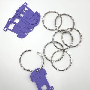 May include: An overhead shot shows a collection of silver metal binder rings and several purple die-cut shapes. The shapes resemble small, stylized houses or buildings. The rings are open and closed, and the shapes are scattered on a white surface.