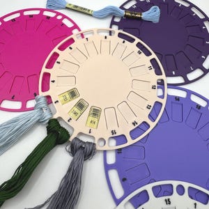 May include: A collection of circular plastic embroidery floss organizers in pink, purple, and cream. Each organizer features numbered slots for thread management. Three skeins of embroidery thread in light blue, dark green, and gray are attached.