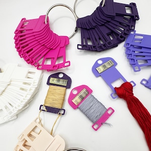 May include: A collection of colorful thread spools and color swatches. The spools are attached to pink, purple, and lavender tags with barcodes. The color swatches are in pink, purple, white, and beige, attached to metal rings.