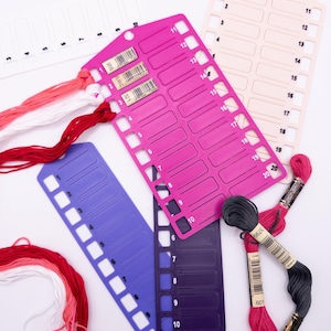 May include: A collection of embroidery floss and thread organizers in various colors. The image shows pink, purple, and white plastic thread organizers with numbered slots. Bundles of red, pink, white, and gray embroidery floss are also visible.