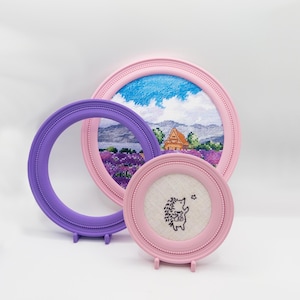 May include: Three circular frames containing embroidery. One large pink frame holds a landscape with a house and lavender fields. A smaller pink frame features a hedgehog embroidery on white fabric. An empty purple frame stands to the left.