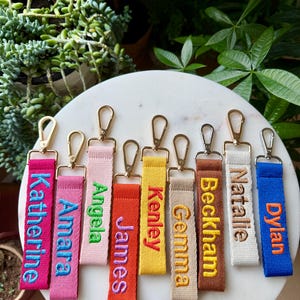May include: A collection of eight fabric keychains in various colors including hot pink, light pink, orange, yellow, beige, brown, cream, and blue. Each keychain features a gold or silver metal lobster clasp and embroidered names.