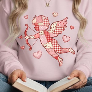 Puede incluir: Sudadera rosa claro con un diseño de Cupido en patchwork, con arco y flecha y motivos de corazón. El Cupido tiene alas y ropa con diseños rosas y rojos. Se ve un collar con la letra "M".