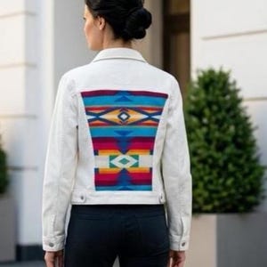 May include: A white denim jacket with a colorful, geometric patterned back panel. The pattern features shades of blue, red, yellow, and green. The jacket has a classic collar, button closures, and long sleeves.