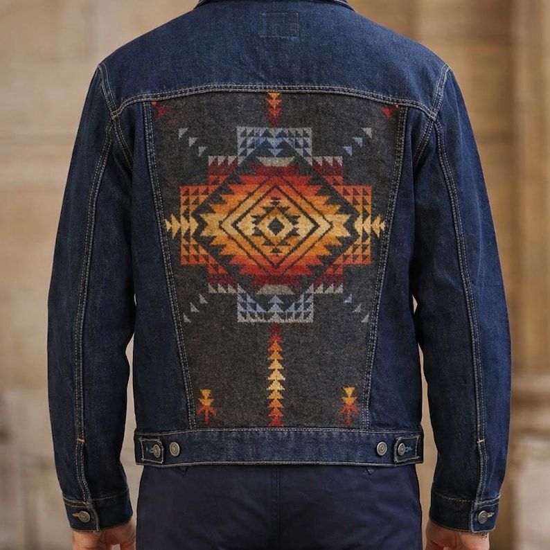 May include: A dark wash denim jacket with a patterned back panel. The panel features a geometric design in shades of orange, yellow, red, and blue on a gray background. The jacket has a classic collar, button closures, and pockets.