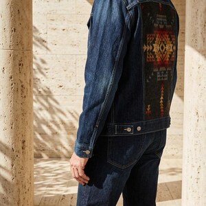 May include: A dark wash denim jacket with a decorative patterned back panel. The back panel features a geometric design in shades of red, orange, and yellow on a dark gray background. The jacket is paired with matching dark wash denim jeans.