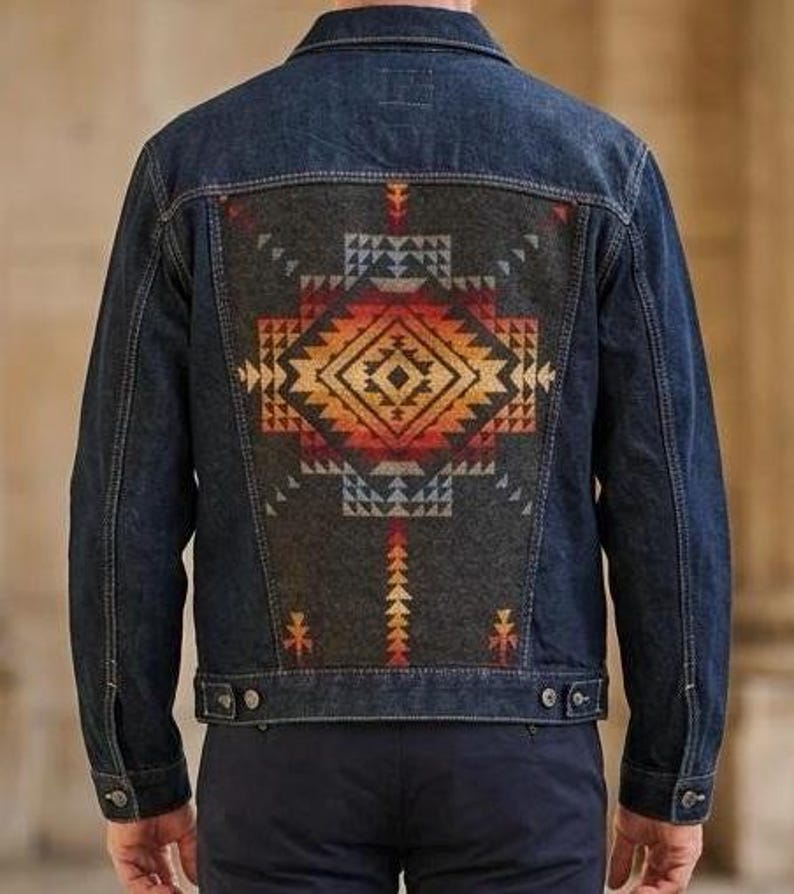 May include: A dark wash denim jacket with a decorative patterned back panel. The panel features a geometric design in shades of orange, red, and blue on a gray background. The jacket has button closures and pockets.