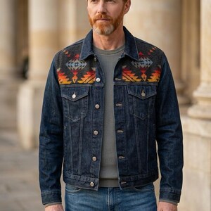 May include: A dark wash denim jacket with a patterned yoke in shades of gray, orange, and yellow. The jacket has a button-up front, chest pockets, and a classic collar. The model is wearing a gray sweater and blue jeans.