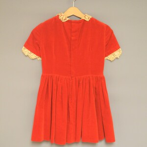 Pu&ograve; includere: Un abito vintage in velluto rosso con maniche corte e gonna a pieghe. L'abito presenta un bordo in pizzo color crema intorno al colletto e ai bordi delle maniche. Appeso a una gruccia di legno su uno sfondo grigio neutro.
