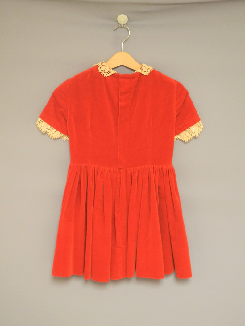 Pu&ograve; includere: Un abito vintage in velluto rosso con maniche corte e gonna a pieghe. L'abito presenta un bordo in pizzo color crema intorno al colletto e ai bordi delle maniche. Appeso a una gruccia di legno su uno sfondo grigio neutro.