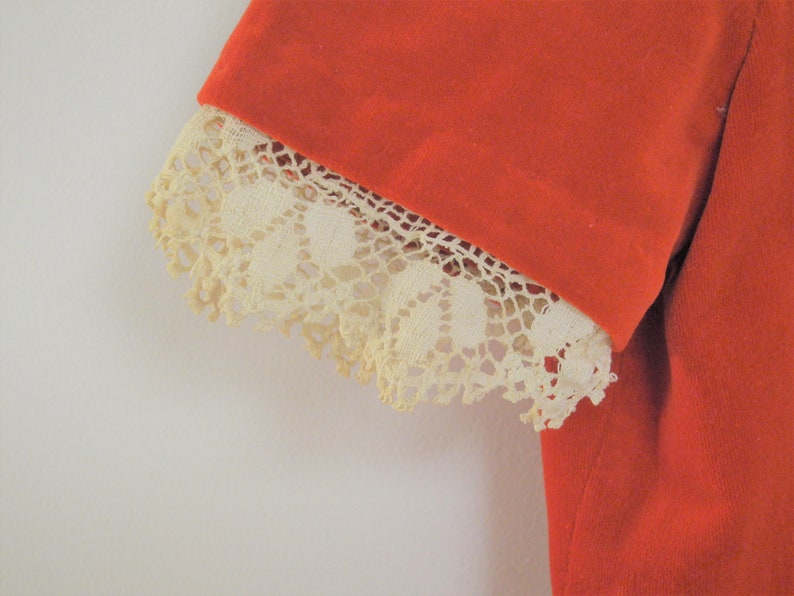 Pu&ograve; includere: Primo piano di una manica di un capo d'abbigliamento. La manica &egrave; di un vibrante colore arancione, con un bordo in pizzo color crema sul polsino. Il pizzo ha un design delicato e traforato.