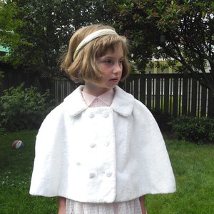 May include: A white, double-breasted cape with a rounded collar. The cape has three sets of two buttons. The person is wearing a pink collared shirt and a white headband. The background is a green lawn and trees.