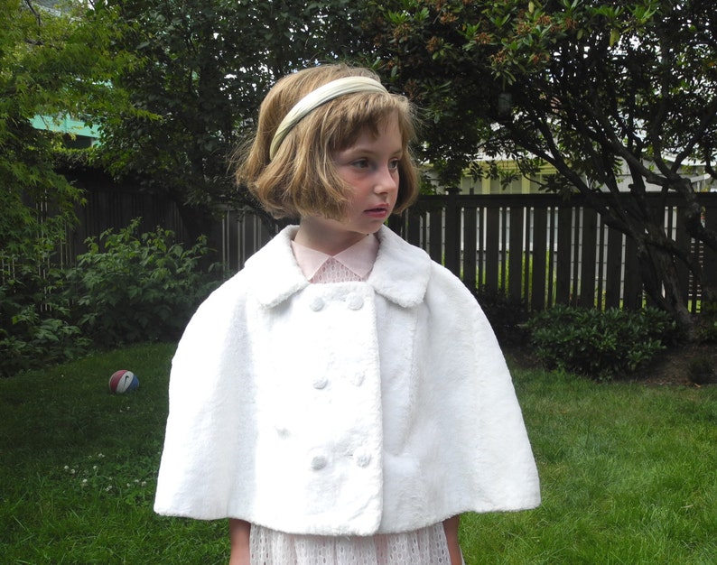May include: A white, double-breasted cape with a rounded collar. The cape has three sets of two buttons. The person is wearing a pink collared shirt and a white headband. The background is a green lawn and trees.