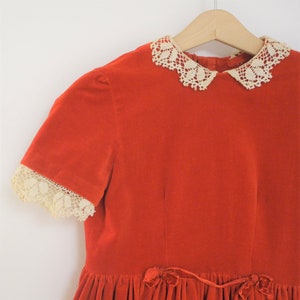 Pu&ograve; includere: Un abito vintage in velluto rosso con colletto e bordi delle maniche in pizzo color crema. L'abito presenta maniche corte e una gonna a balze, con un fiocco in velluto in vita. &Egrave; appeso a una gruccia di legno.