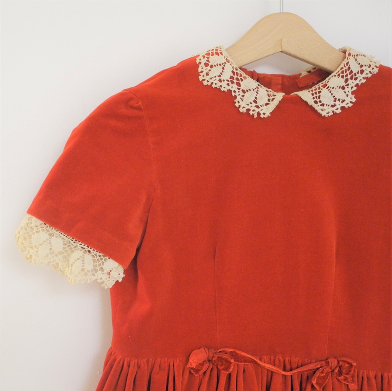 Pu&ograve; includere: Un abito vintage in velluto rosso con colletto e bordi delle maniche in pizzo color crema. L'abito presenta maniche corte e una gonna a balze, con un fiocco in velluto in vita. &Egrave; appeso a una gruccia di legno.