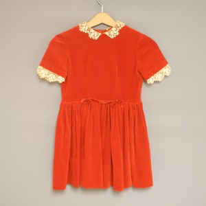 Pu&ograve; includere: Un vestito vintage in velluto rosso con colletto e maniche corte in pizzo color crema. L'abito presenta una gonna a pieghe e un piccolo fiocco in vita. Appeso a una gruccia di legno.