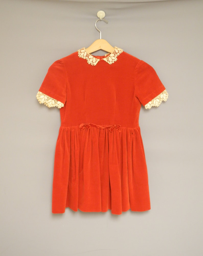 Pu&ograve; includere: Un vestito vintage in velluto rosso con colletto e maniche corte in pizzo color crema. L'abito presenta una gonna a pieghe e un piccolo fiocco in vita. Appeso a una gruccia di legno.
