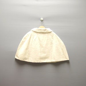 May include: Cream-colored cape with a rounded collar, hanging on a wooden hanger. The cape is made of a textured fabric and is displayed against a gray background. The garment is a classic outerwear piece.