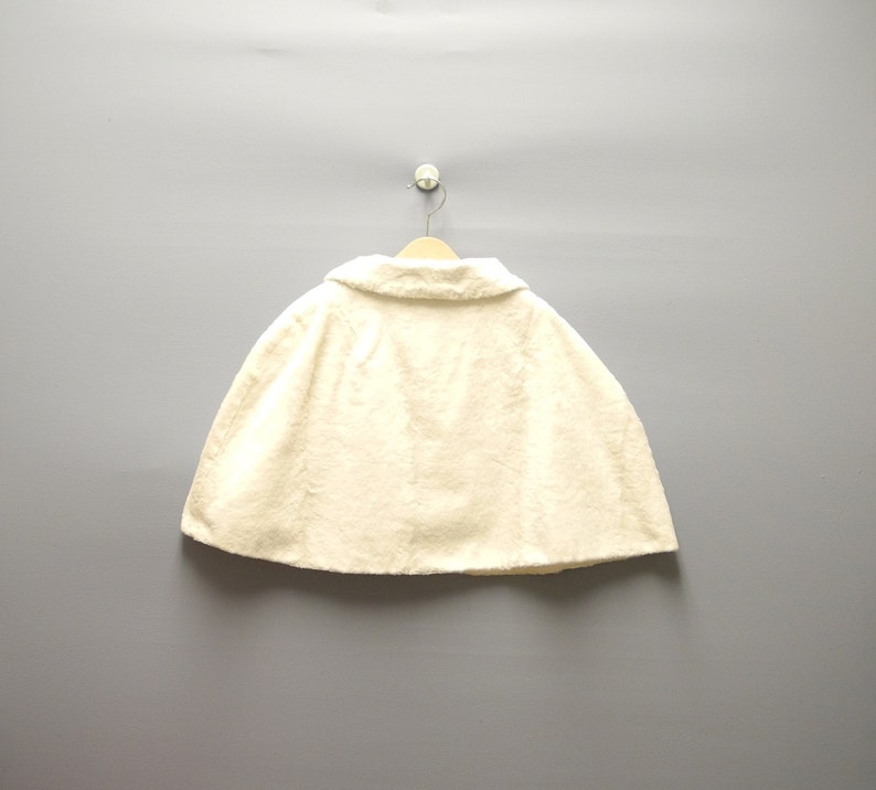 May include: Cream-colored cape with a rounded collar, hanging on a wooden hanger. The cape is made of a textured fabric and is displayed against a gray background. The garment is a classic outerwear piece.