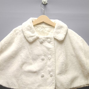 May include: A cream-colored faux fur cape with a rounded collar and double-breasted button closure. The cape is hanging on a wooden hanger against a gray background. The soft, fluffy texture of the cape is visible.