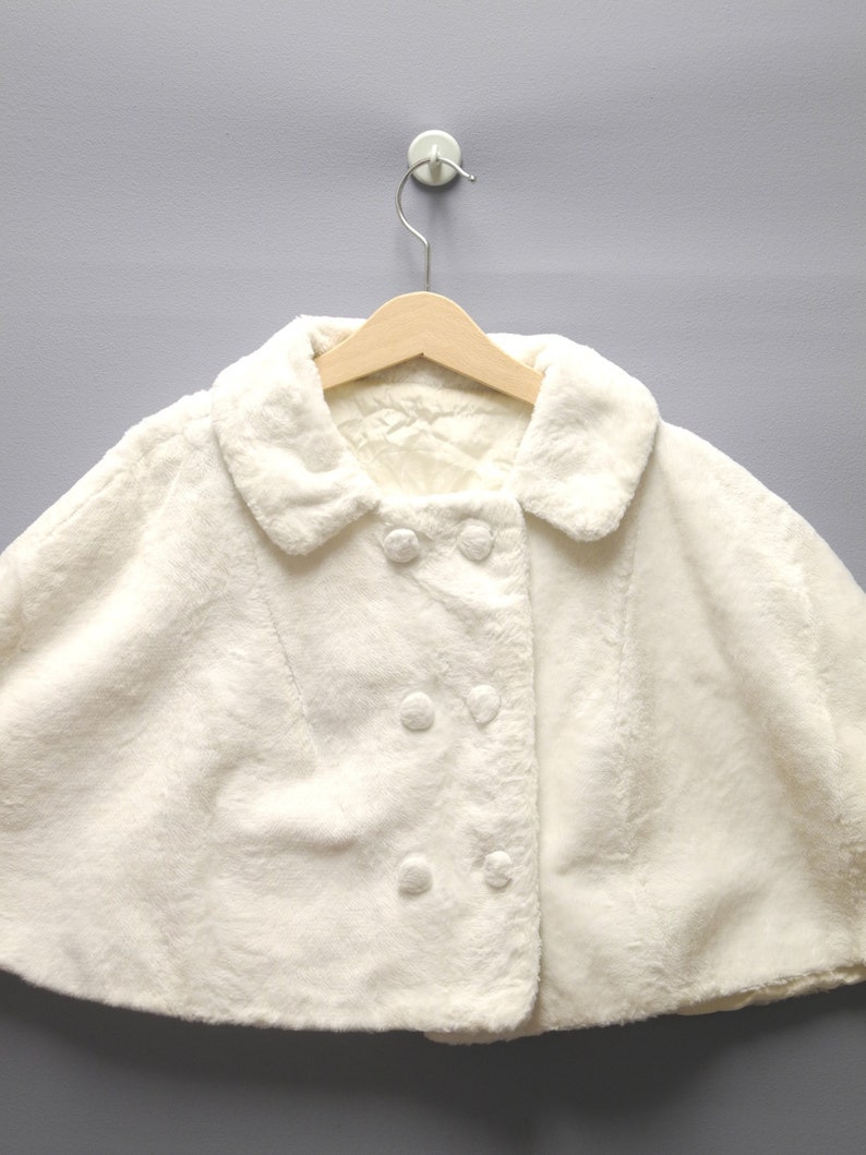 May include: A cream-colored faux fur cape with a rounded collar and double-breasted button closure. The cape is hanging on a wooden hanger against a gray background. The soft, fluffy texture of the cape is visible.