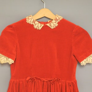 Pu&ograve; includere: Un vestito vintage in velluto rosso con colletto e bordi delle maniche in pizzo color crema. L'abito presenta maniche corte e una gonna a pieghe, con un dettaglio a fiocco in vita. Appeso a una gruccia di legno.