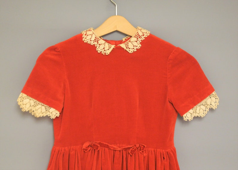 Pu&ograve; includere: Un vestito vintage in velluto rosso con colletto e bordi delle maniche in pizzo color crema. L'abito presenta maniche corte e una gonna a pieghe, con un dettaglio a fiocco in vita. Appeso a una gruccia di legno.