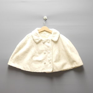 May include: Cream-colored cape with a rounded collar and button closure. The cape is made of a soft, textured fabric and hangs on a wooden hanger against a gray background. The cape has a double-breasted button design.