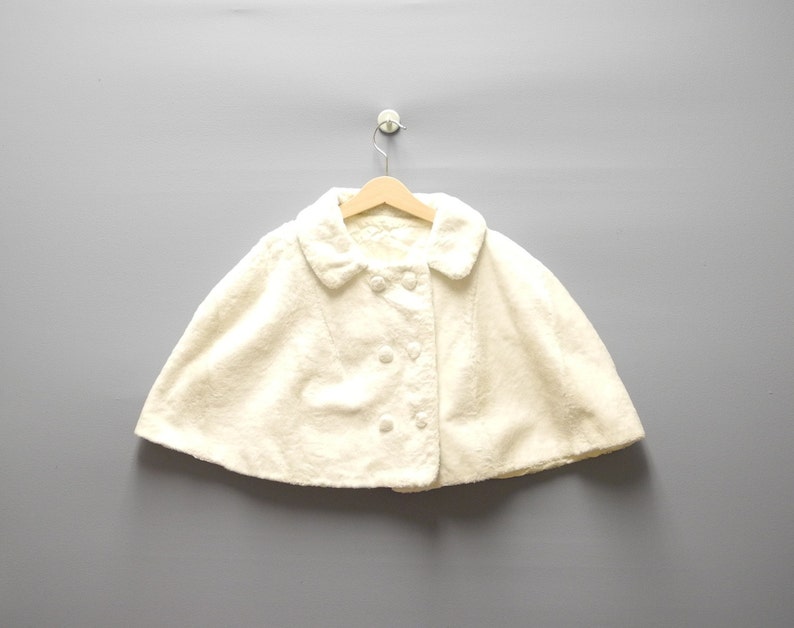 May include: Cream-colored cape with a rounded collar and button closure. The cape is made of a soft, textured fabric and hangs on a wooden hanger against a gray background. The cape has a double-breasted button design.