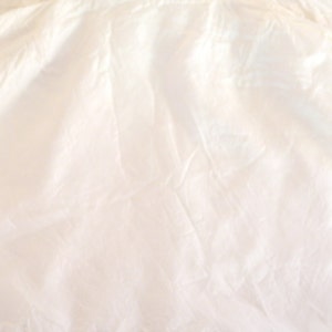 May include: A close-up shot of a cream-colored fabric with soft folds and subtle shadows. The fabric appears to be lightweight, possibly silk or a similar material, with a slightly translucent quality. The overall texture is smooth and flowing.