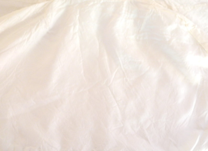May include: A close-up shot of a cream-colored fabric with soft folds and subtle shadows. The fabric appears to be lightweight, possibly silk or a similar material, with a slightly translucent quality. The overall texture is smooth and flowing.