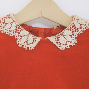 Pu&ograve; includere: Un abito rosso brillante con un colletto in pizzo color crema, appeso a una gruccia di legno. Il colletto presenta un delicato motivo traforato. L'abito &egrave; realizzato in un tessuto morbido e strutturato.