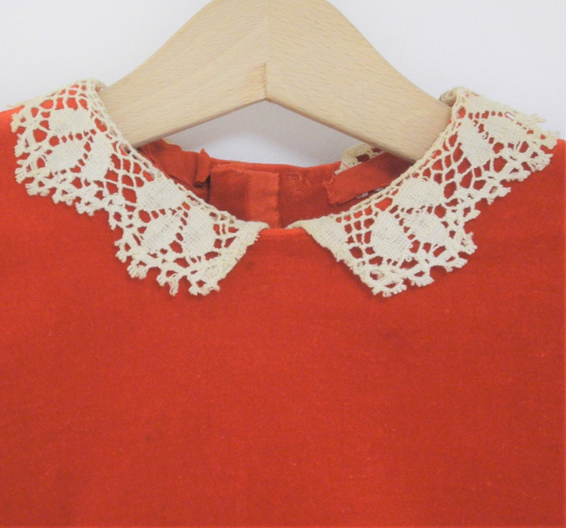 Pu&ograve; includere: Un abito rosso brillante con un colletto in pizzo color crema, appeso a una gruccia di legno. Il colletto presenta un delicato motivo traforato. L'abito &egrave; realizzato in un tessuto morbido e strutturato.