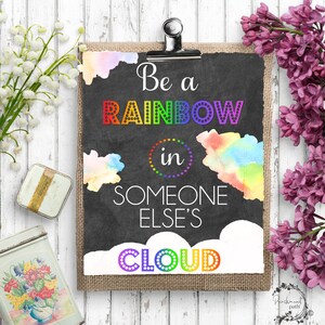 Könnte beinhalten: Ein Schild aus Schiefer mit einem Aquarellhintergrund und dem Text "Be a rainbow in someone else's cloud". Der Text ist in Regenbogenfarben gehalten und der Hintergrund ist ein hellblauer, rosa und gelber Aquarell-Wasch.
