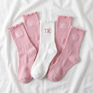 May include: Five pairs of socks, four pink and one white, with ruffled tops and initial embroidery. The pink socks have white initials: XD, WH, KU, and BV. The white sock has the initials TK. All socks have grip dots on the soles.