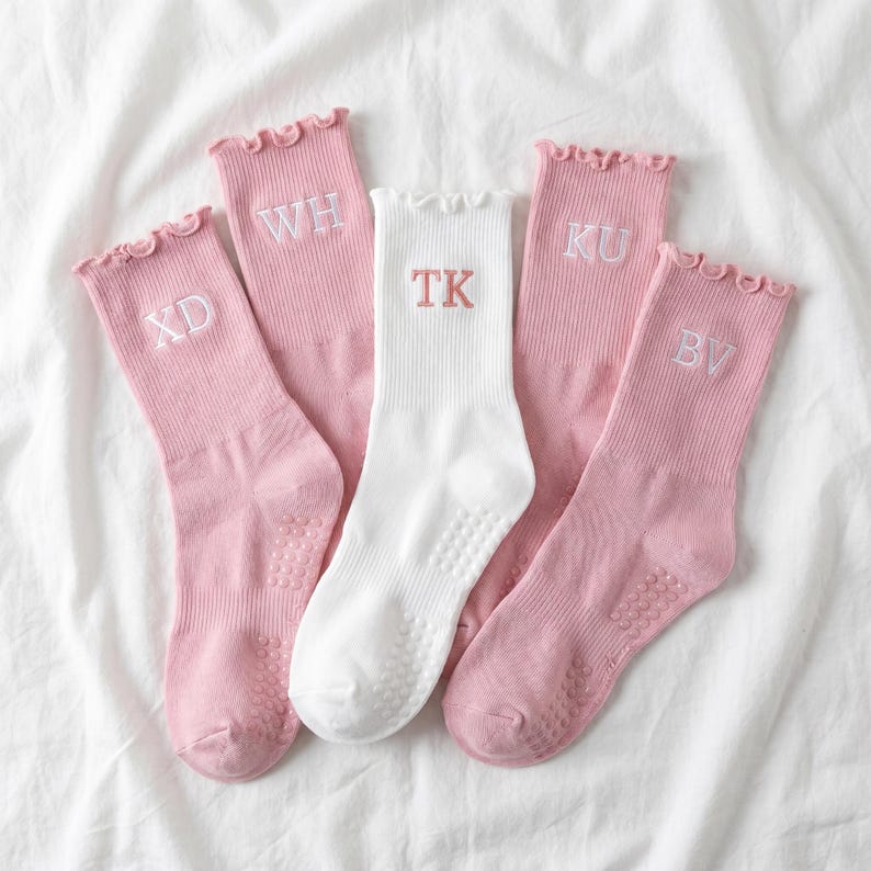 May include: Five pairs of socks, four pink and one white, with ruffled tops and initial embroidery. The pink socks have white initials: XD, WH, KU, and BV. The white sock has the initials TK. All socks have grip dots on the soles.