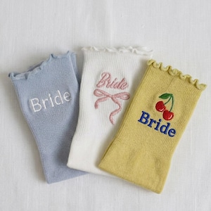 May include: Three pairs of ruffled-top socks in pastel colors. One light blue sock has "Bride" embroidered in white. A white sock features "Bride" with a pink bow. A yellow sock has "Bride" and cherry embroidery.