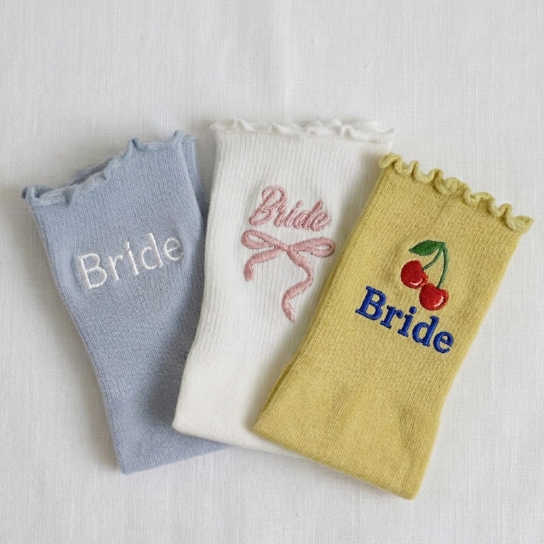 May include: Three pairs of ruffled-top socks in pastel colors. One light blue sock has "Bride" embroidered in white. A white sock features "Bride" with a pink bow. A yellow sock has "Bride" and cherry embroidery.