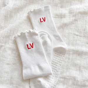 May include: Two pairs of white socks with red embroidered "LV" initials. The socks have a ribbed texture and ruffled tops. One pair has a non-slip grip on the sole.