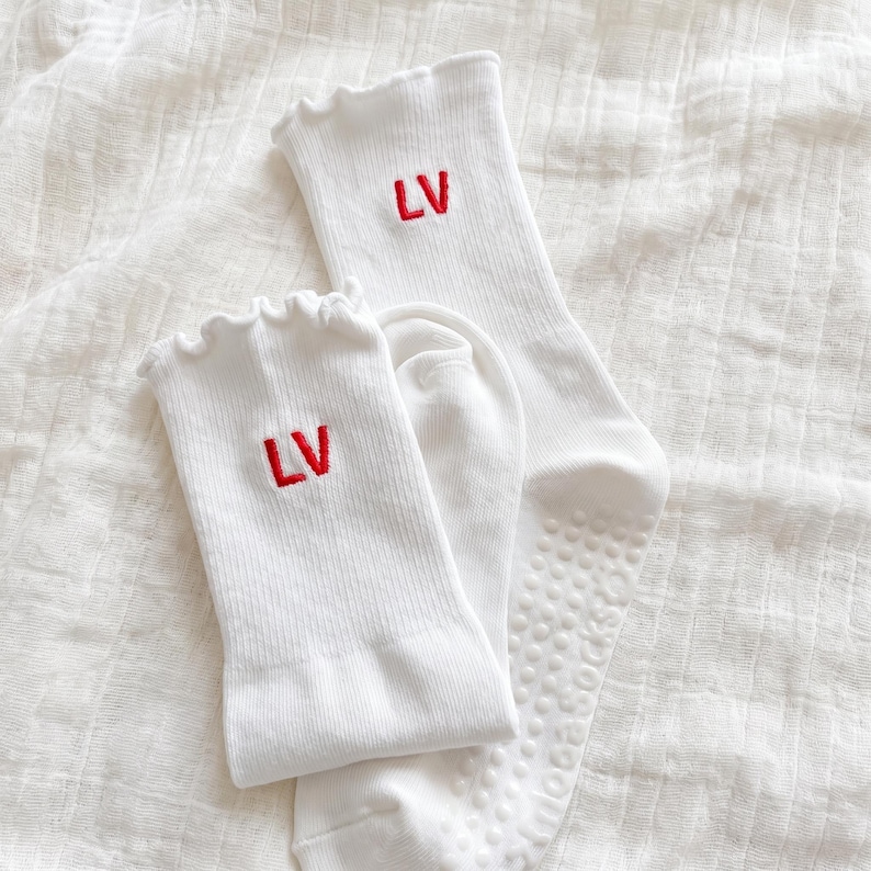 May include: Two pairs of white socks with red embroidered "LV" initials. The socks have a ribbed texture and ruffled tops. One pair has a non-slip grip on the sole.
