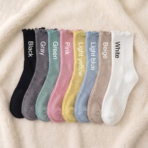 May include: A collection of nine pairs of socks in various colors, including black, gray, green, pink, light yellow, light blue, beige, and white. Each sock has a ruffled top and the color name printed on it.