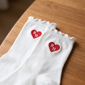 May include: A pair of white socks with a ruffled top edge. Each sock features a red heart with the embroidered letters "RH". The socks are made of a soft, ribbed material.
