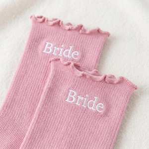May include: Two pairs of pink ribbed socks with a ruffled top edge. The word "Bride" is embroidered in white on each sock. The socks are made of a soft, textured material.