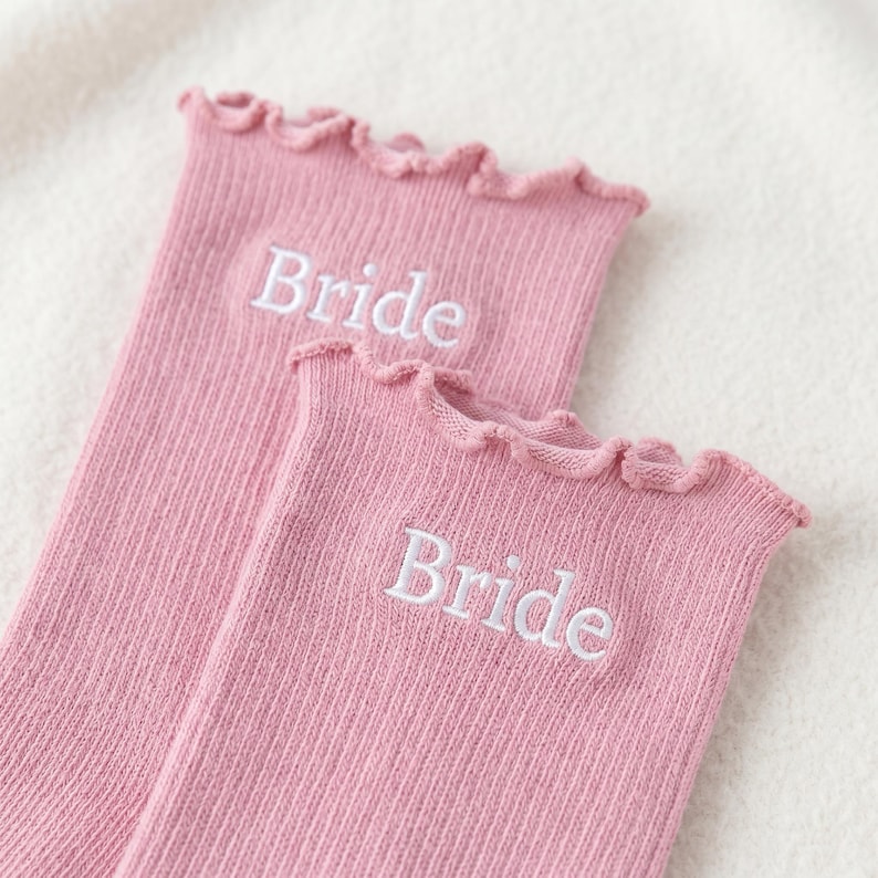 May include: Two pairs of pink ribbed socks with a ruffled top edge. The word "Bride" is embroidered in white on each sock. The socks are made of a soft, textured material.