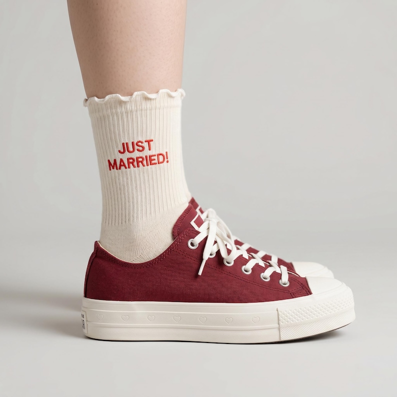 May include: Maroon canvas sneakers with white laces and a white sole featuring heart-shaped cutouts. Cream-colored socks with a ruffled top and the text "JUST MARRIED!" in red are visible.