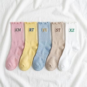 May include: Five pairs of colorful socks arranged on a white surface. Each sock is a different pastel color: pink, yellow, blue, tan, and white. Each sock has a different embroidered letter combination: KM, RT, YU, ST, and XZ.